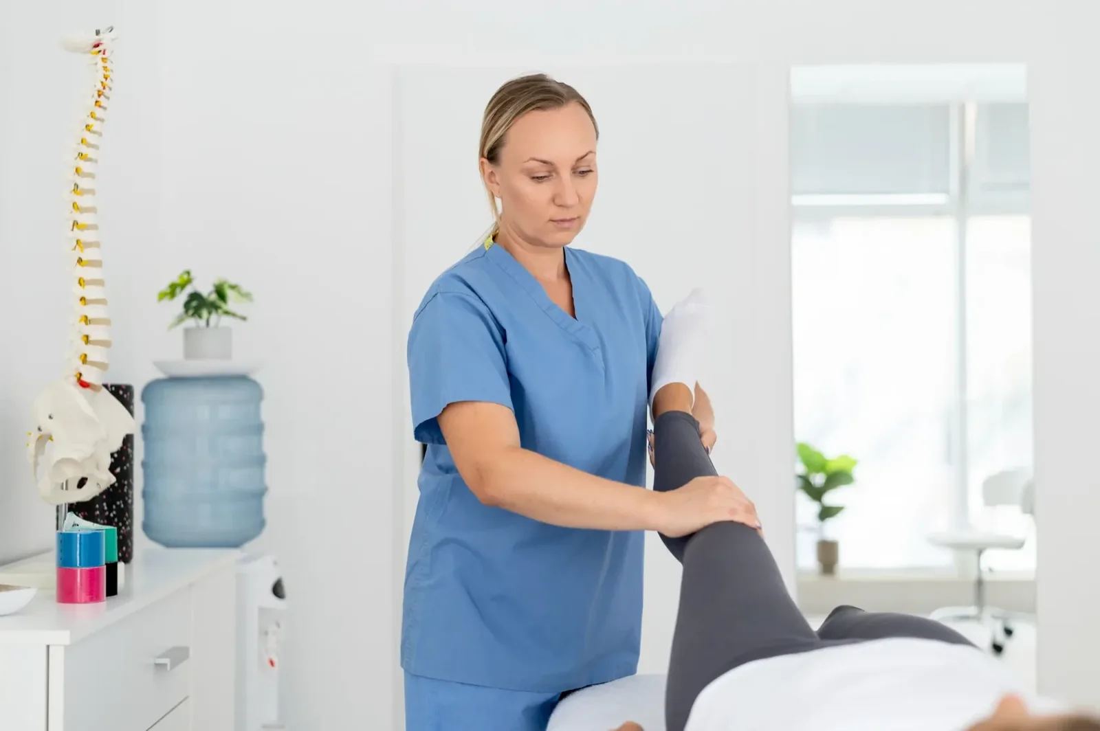 A terapeuta a kezelőágyon fekvő páciens lábát kezeli, mellette gerincmodell (illusztráció)